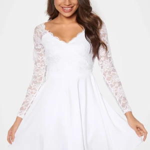 Vit spetsklänning med lång ärm - Superfin vit klänning med v-ringad spets upptill och långa spetsärmar. Kjolen är slät och har en lätt utställd form. Perfekt för student, konfirmation eller midsommar! Dragkedja i ryggen och spetsdetaljer med blommor. Endast använd en gång på min konfirmation! 