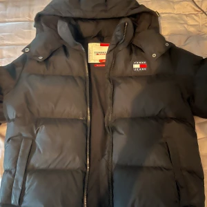 Svart pufferjacka från Tommy Jeans - Säljer en svart pufferjacka från Tommy hilfiger med huva och dragkedja. Perfekt för kalla dagar och har en clean, stilren look. Den är jättevarmt och bekväm vilket gör den perfekt för vinter Jackan är även i helt perfekt skick. Pris kan justeras vid snabb affär