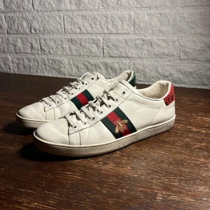 Gucci Ace - Storlek 40 Skick 8/10 (liten defekt på vänster sko). Gucci Ace sneakers i vitt skinn med klassiska gröna och röda ränder på sidorna samt broderad guldfärgad bi. Baksidan har metallicröd och grön detalj. Platt sula med unikt mönster undertill och snörning framtill. Perfekta för dig som vill sticka ut med lyxig streetstyle.