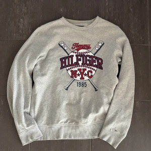 Grå sweatshirt Tommy Hilfiger NYC - Sällsynt sweatshirt som ej säljs längre eller går att hitta! Grå sweatshirt från Tommy Hilfiger med stort NYC-baseballtryck framtill i rött, blått och vitt. Klassisk rund hals, ribbade muddar och Hilfiger-logga på ärmen. Perfekt för dig som gillar sportig och avslappnad stil.