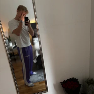 Palm Angels lila trackpants - Säljer ett par lila trackpants från Palm Angels med vita sidoränder och elastisk midja. Byxorna har logga tryckt på ena benet och är tillverkade i ett glansigt syntetmaterial. Perfekta för en sportig och trendig look.