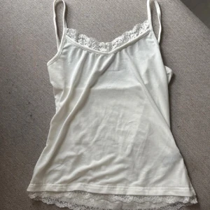 Vit linnetopp med spetskant - Säljer en söt vit linnetopp med tunna axelband och dekorativ spetskant både vid halsringningen och längst ner. Toppen är mjuk och skön, perfekt för varma dagar eller som layering-piece. Enkel och stilren design som funkar till många looks.