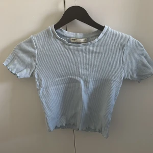 Ljusblå ribbad topp Pull&Bear - Säljer en ljusblå ribbad topp från Pull&Bear med korta ärmar och rund hals. Toppen har en croppad passform och vågig kant nertill och vid ärmsluten. Perfekt för dig som gillar en enkel men trendig stil.