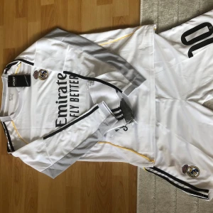 Real Madrid långärmad fotbollströja Mbappé - Säljer en långärmad vit Real Madrid fotbollströja med Mbappé och nummer 10 på ryggen. Tröjan har gråa och svarta detaljer, gula inslag och klubbmärke på bröstet. Ingår matchande shorts och strumpor. Perfekt för dig som älskar fotboll och vill sticka ut på planen.