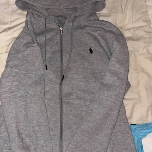 Grå hoodie från Polo Ralph Lauren - Säljer en stilren grå hoodie från Polo Ralph Lauren med dragkedja och huva. Tröjan har det klassiska broderade logotypen på bröstet och justerbara snören i huvan. Perfekt för chill dagar eller när du vill ha en clean look.