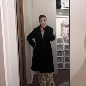 Svart kappa från KappAhl, stl M - Stilren svart kappa från KappAhl i storlek M. Klassisk rak modell med krage, en knapp framtill och diskreta fickor på sidorna. Perfekt för dig som gillar en clean och tidlös look. Materialet är mjukt och känns lyxigt.