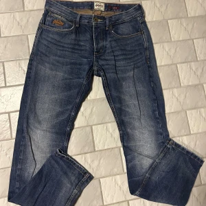 Blå jeans från Superdry i bomull - Snygga blå jeans från Superdry med klassisk femficksdesign och diskreta slitningar. Jeansen är tillverkade i 100% bomull och har raka ben med normal passform. Perfekta för en avslappnad och stilren look.