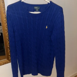 Blå stickad tröja från Ralph Lauren - Säljer en klassisk blå stickad tröja från Ralph Lauren med kabelstickat mönster och gul broderad logga på bröstet. Tröjan har rund halsringning och långa ärmar, perfekt för en stilren och chill look. Modellen är 186 cm lång. Storleken är Junior L 14-16 men sitter som en Vuxen S.