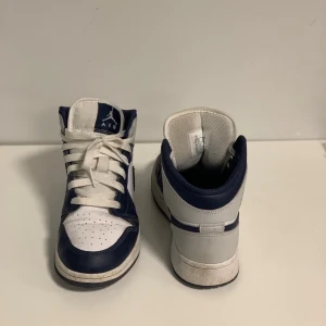Nike Air Jordan 1 Mid Navy/Vit - Säljer ett par Nike Air Jordan 1 Mid i marinblått och vitt. Klassisk siluett med snörning, perforerad tå och ikoniska Jordan-loggor på sidan och plösen. Ovandel i skinn och syntet, med vit sula och mörkblå detaljer. Perfekta för dig som gillar streetstyle och sneakers med attityd.