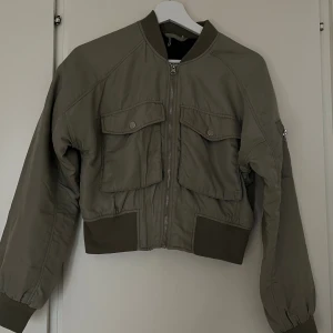 Olivgrön croppad bomberjacka från H&M - Olivgrön bomberjacka med croppad passform, stora fickor framtill och dragkedja. Jackan har ribbade muddar vid ärmslut, krage och midja. Något fläckig från användning.