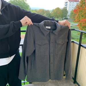 Grå overshirt från These Glory Days - Snygg grå overshirt från These Glory Days i ullmix. Klassisk rak modell med krage, knäppning framtill och två bröstfickor med lock. Perfekt lager-på-lager-plagg med stilren vibe och mjukt material.