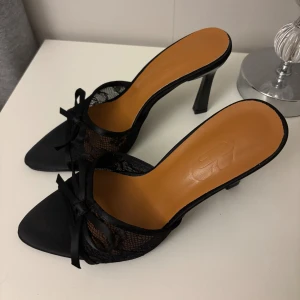 Svarta spetsiga pumps med rosett - Säljer ett par svarta pumps med spetsig tå och hög klack. Skorna har transparent spetsdetalj på sidorna och en söt rosett framtill. Snygg och elegant design med öppen häl och platt sula framtill. Perfekta för dig som gillar en chic och trendig look.