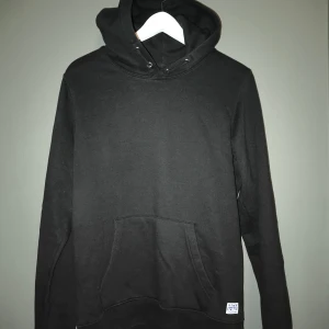 Svart hoodie från Jack & Jones - Svart hoodie från Jack & Jones med klassisk känguruficka och huva med snörning. Tröjan är i mjuk bomull och har en enkel, stilren design som passar perfekt till jeans eller joggers.