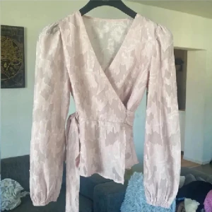 Ljusrosa omlottblus med knytband - Supersöt ljusrosa omlottblus med subtilt blommönster och v-ringning. Blusen har långa puffärmar med resår vid handlederna och knytband i midjan för en snygg siluett. Materialet är lätt och har en diskret struktur som ger en lyxig känsla. Endast swish skickas spårbart 59kr