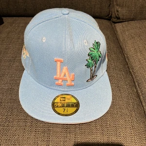 Blå LA Dodgers keps New Era 59FIFTY - Snygg ljusblå LA Dodgers keps från New Era 59FIFTY med broderade palmer och jubileumsmärke på sidan. Kepsen har rosa LA-logga framtill och rosa underskärm. Perfekt för dig som vill vara chill med en unik och färgglad look.