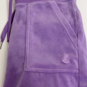 Lila mjukisbyxor Juicy Couture XS - Lila mjukisbyxor från Juicy Couture i supermjuk velour med bred resår i midjan och snörning. Byxorna har stora fickor fram och bak, varav bakfickan har tryckknappar med logga. Perfekta för en chill och trendig look.