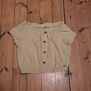 Gul offshoulder topp från Hatton Republic - Söt ljusgul offshoulder topp från Hatton Republic med korta ärmar och knappar framtill. Toppen har resår vid halsringningen och nederkanten för en avslappnad passform. Perfekt för varma dagar och enkel att matcha med jeans eller kjol.