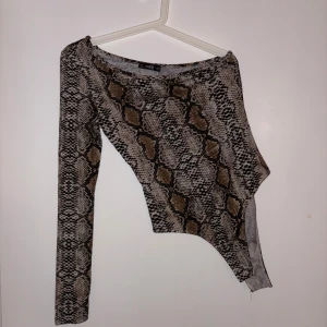 Oneshoulder topp med ormmönster - Säljer en oneshoulder topp med ormmönster i beige, brunt och svart. Toppen har lång ärm på ena sidan och är ärmlös på andra, vilket ger en cool asymmetrisk look. Tillverkad i stretchigt syntetmaterial som sitter tight och markerar figuren.