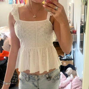 Vit blus med volang och öppen rygg - Supersöt vit blus med broderade detaljer, volangaxlar och öppen rygg med knytband. Luftig och lätt med romantisk känsla, perfekt till jeans eller kjol. Den har fyrkantig ringning och är ärmlös med breda axelband.