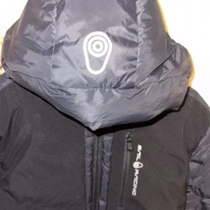 Svart dunjacka från Sail Racing - Säljer en svart dunjacka från Sail Racing med huva och dragkedja. Jackan har en bröstficka med dragkedja och logga på bröstet samt patch på ärmen. Fylld med ansvarsfullt framtaget dun (500 FP) och har en sportig look. Perfekt för kalla dagar.