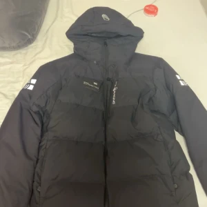 Svart dunjacka från Antarctic Expedition - Svart dunjacka med huva från Antarctic Expedition. Jackan har en quiltad design, dragkedja framtill och tryckta loggor på ärmar och bröst. Fylld med 500 FP ansvarsfullt framtaget dun, perfekt för kalla dagar. Snygg och funktionell med en clean look.