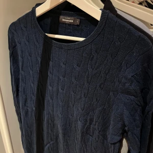 Mörkblå kabelstickad tröja Dressmann - Kabelstickad mörkblå tröja från Dressmann i storlek S. Tröjan har rund halsringning och långa ärmar, perfekt för lager-på-lager. Klassisk och stilren design som funkar till många olika outfits.