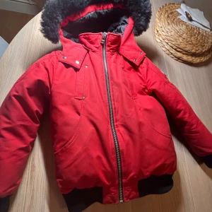 Röd dunjacka Moose Knuckles M - Röd dunjacka från Moose Knuckles med svart fuskpäls på huvan, silverfärgade detaljer och dragkedja framtill. Jackan har svarta muddar, innerfoder i svart och röd, samt logga på ärmen. Perfekt för kalla dagar och riktigt snygg streetstil.