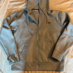 Lacoste hoodie  - Snygg ljusblå hoodie från Lacoste med dragkedja och klassisk krokodillogga på bröstet. Tröjan har huva med snörning, två fickor fram och är tillverkad i mjuk bomull. Perfekt för chill dagar eller när du vill ha en clean look. 7/10 skick passar om du är runt 180