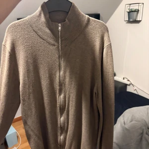 Beige kofta med dragkedja från Lindbergh - Snygg beige kofta från Lindbergh med hög krage och hel dragkedja framtill. Tröjan är stickad och har långärmad design, perfekt för lager-på-lager. Passformen är slim och materialet känns mjukt och bekvämt.