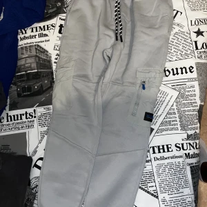 Grå cargo mjukisbyxor från McKenzie - Grå mjukisbyxor från McKenzie med svarta snören i midjan och praktiska cargofickor med dragkedja på benen. Byxorna har en loose fit och är tillverkade i mjukt bomullsmaterial, perfekta för chill dagar. Snygga detaljer med logga på benet och kontrastfärgade snören.