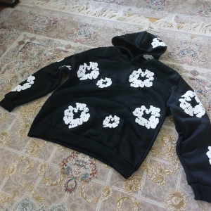 Svart hoodie med vita blommiga tryck Denim tears - Cool svart hoodie med stora vita blommiga tryck över hela tröjan, även på huvan. Tröjan har klassisk känguruficka och huva. Perfekt för dig som vill sticka ut med en unik och trendig stil.