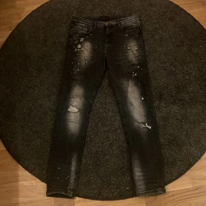 Svarta jeans med slitningar - Säljer ett par svarta  jeans med coola slitningar och färgstänk-detaljer. Jeansen har en klassisk femficksdesign. Perfekta för dig som gillar en edgy streetstil. Materialet är jeans och färgen är svart med ljusa tvättade partier.