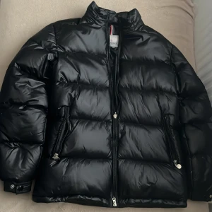 Svart Moncler pufferjacka - Säljer en svart pufferjacka från Moncler med glansig finish och klassisk Moncler-logga på ärmen. Jackan har hög krage, två dragkedjefickor och hel dragkedja framtill. Perfekt för kalla dagar och riktigt snygg till streetwear-stilen.
