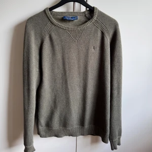 Olivgrön stickad tröja från Polo Ralph Lauren - Snygg olivgrön stickad tröja från Polo Ralph Lauren med diskret broderad logga på bröstet. Tröjan har rund halsringning, ribbade muddar och lång ärm. Perfekt nu när det börjar bli lite kyligare ute.