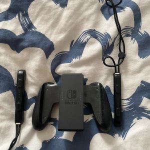 Nintendo Switch Joy-Con Grip och Joy-Con Remmar - Nintendo Switch Joy-Con Grip och två Joy-Con Remmar i svart. Passar perfekt för att spela med Joy-Cons som en traditionell handkontroll. Remmarna har handledsband för extra säkerhet. Mindre repor och normalt bruksslitage syns, men allt är i gott skick.