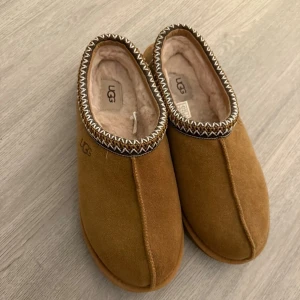 Bruna UGG tofflor mocka - Mysiga bruna tofflor från UGG i mocka med mjukt foder på insidan. Dekorativt mönster runt öppningen och robust gummisula. Perfekta för kalla dagar när du vill ha det extra bekvämt. Använda endast 1gång. Nypris 1700kr