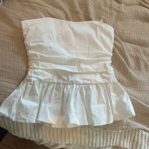 Vit volang tubtopp från Zara - Säljer en vit tubtopp från Zara med volang nedtill och lätt rynkade detaljer i midjan. Toppen är axelbandslös och har en stilren, feminin look som passar perfekt till sommaren. Materialet är mjukt och känns skönt mot huden.