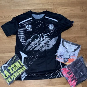 Jul x 90 Minute kortärmad t-shirt - Säljer en cool t-shirt från Jul x 90 Minute med grafiska tryck, alienmotiv och text som 'La Zone en Personne' och 'Toto & Ninietta'. T-shirten är kortärmad, har rund hals och är gjord i lätt polyester. Färgerna är svart, vit, gul och orange med detaljer i grått.