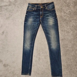 Mörkblå slim fitt  jeans från Nudie - Snygga mörkblå slim fitt  jeans från Nudie Jeans med slitningar och kontrastsömmar. Klassisk femficksmodell med läderpatch bak och tydliga whiskers på knä och lår. Perfekt passform och riktigt sköna att bära.