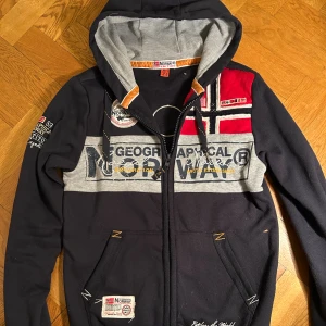 Geographical Norway hoodie med tryck - Mörkblå hoodie från Geographical Norway med grå detaljer, broderade och tryckta loggor samt norska flaggan på bröstet. Tröjan har huva, dragkedja, fickor och ribbade muddar. Sjukt bra passform i storlek S.