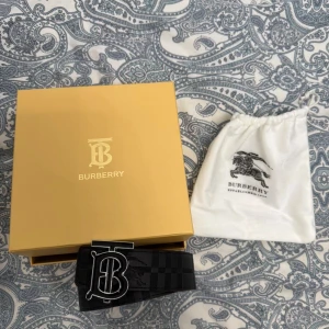 Svart Burberry bälte med rutigt mönster - Snyggt svart bälte från Burberry med klassiskt rutigt mönster och stor metallspänne med Burberrys ikoniska logotyp. Bältet är tillverkat i skinn och har en lyxig känsla, perfekt för att lyfta din outfit med en exklusiv touch. Du kan även byta sida mellan den svarta och den normala burberry loggan.