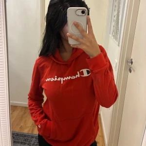 Röd hoodie från Champion 162/167 - Röd hoodie från Champion i storlek 162/167 med vit logga över bröstet. Klassisk känguruficka framtill, huva och långa ärmar. Perfekt för chill dagar eller när du vill ha en sportig vibe. Mjuk och skön bomullskänsla. Sitter som storlek S/XS