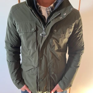 Grön fieldjacket från Barbour - Snygg mörkgrön fieldjacket från Barbour med flera stora fickor framtill, hög krage och dragkedja med tryckknappar. Jackan är tillverkad i polyester och har en klassisk, rak passform. Perfekt för kyliga dagar och har en stilren look. Passar S-M