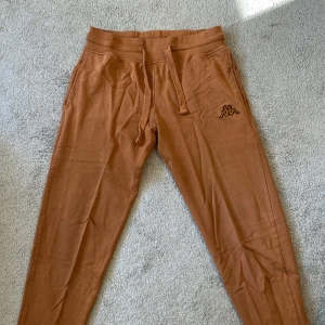 Bruna mjukisbyxor från Kappa - Snygga bruna mjukisbyxor från Kappa med broderad logga på ena benet. Byxorna har dragsko i midjan, ribbade muddar vid benslut och praktiska fickor fram samt en bakficka. Perfekta för chill eller träning.