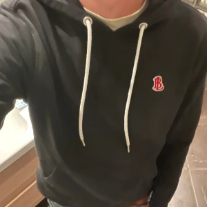 Svart hoodie med vit snörning och brodyr - Svart hoodie med vit snörning och broderad BL-logga i rött och vitt på bröstet. Klassisk känguruficka framtill och huva. Perfekt för chill dagar och enkel att matcha med jeans eller joggers.