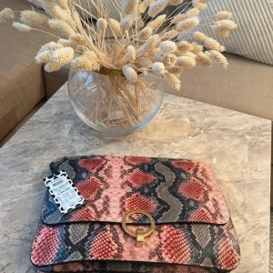 Axelväska i ormskinnsmönster med ringdetalj - Snygg axelväska i syntetmaterial med ormskinnsmönster i rött, svart och grått. Väskan har en avtagbar axelrem och en guldig ringdetalj på locket. Perfekt för dig som vill sticka ut med en trendig accessoar.