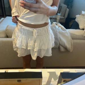 Vit volangkjol från Pull&Bear - Supersöt vit kortkjol från Pull&Bear med volanger och knyt i midjan. Kjolen har en luftig och lätt känsla, perfekt för varma dagar. Den har en feminin och flowy siluett som ger en avslappnad men ändå trendig vibe.