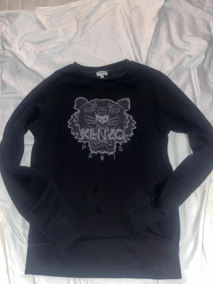 Svart sweatshirt från Kenzo med tiger - Svart sweatshirt från Kenzo i storlek S med ikonisk broderad tiger och Kenzo Paris-logga på bröstet. Tröjan har rund hals, långa ärmar och ribbade muddar. Materialet är mjuk bomull som känns skön mot huden. Perfekt för dig som gillar streetwear och snygga detaljer. 