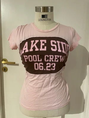 Rosa och brun t-shirt med tryck - T-shirt i ljusrosa med brunt parti över bröstet och texten 'LAKE SIDE POOL CREW 06.23' i rosa bokstäver. Kortärmad modell med figurnära passform och rund halsringning. Tillverkad i mjuk bomull, perfekt för en avslappnad look.
