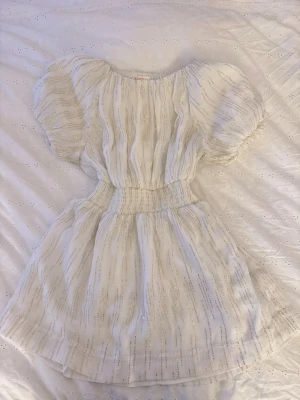 Glittrig nyårs klänning  - Supersöt vit klänning med glittriga ränder och puffiga kortärmar. Klänningen har smock i midjan för en snygg siluett och är tillverkad i ett lätt, glittrigt material. Nytt skick har ej kommit till användning 